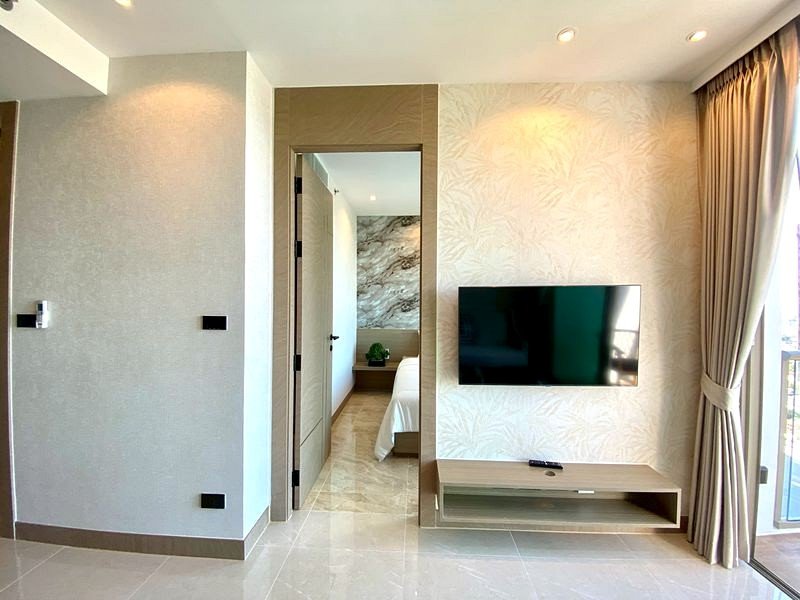 Living area of a modern hotel suite with a wall-mounted TV above a low console, beige wallpaper, and floor-to-ceiling curtains by a balcony door.