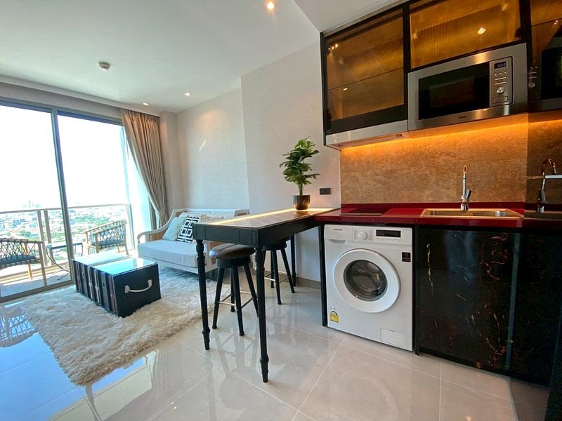 Open-plan living room and kitchen in a modern apartment: white sofa and cream rug beside a black dining table, washing machine under the red countertop, microwave above, and a potted plant on the table with a balcony view through glass doors to the left.