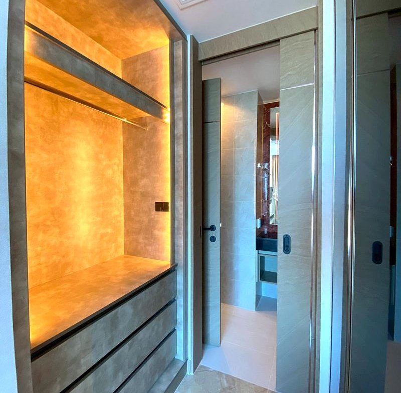 Modern bathroom with a warm-lit built-in storage alcove on the left and a doorway to a marble-tiled bathroom ahead, visible through glass doors.