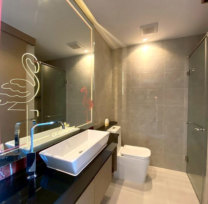 Modern bathroom with a black countertop, white rectangular sink, and a large illuminated mirror featuring a neon flamingo design and 'OCEAN DRIVE' text, plus a toilet and glass shower area.