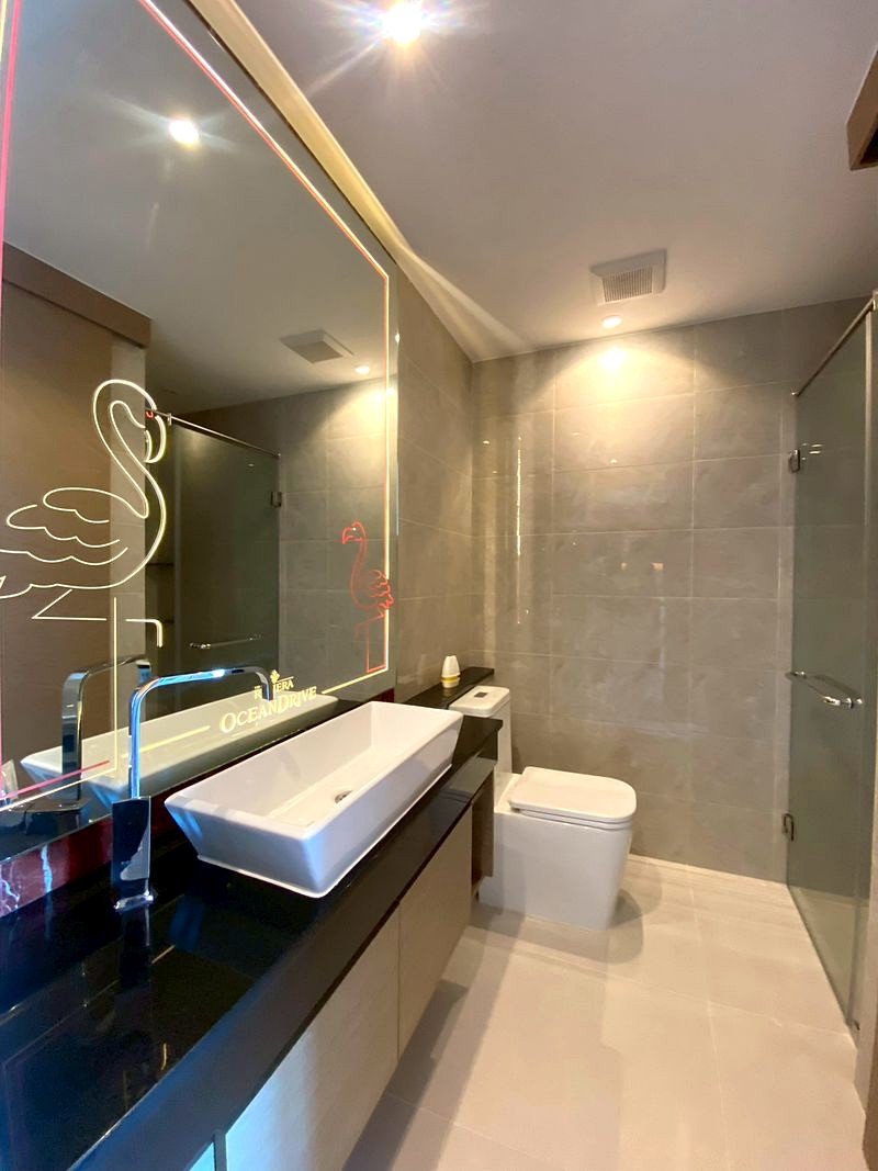 The_Riviera_Ocean_Drive_3214_7 Modern bathroom with a black countertop, white rectangular sink, and a large illuminated mirror featuring a neon flamingo design and 'OCEAN DRIVE' text, plus a toilet and glass shower area.