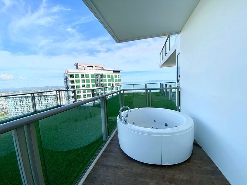 On a high-rise balcony, a round white jacuzzi sits on a wooden deck with glass railing and a distant cityscape under a clear blue sky.