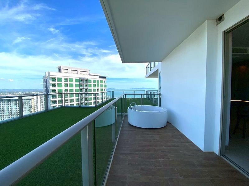 Balcony with a glass railing, a white round hot tub, and wooden decking overlooking the city and sea.