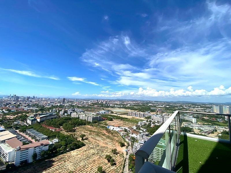 Aerial view from a high-rise balcony with glass railing overlooking a city and open fields under a bright blue sky.