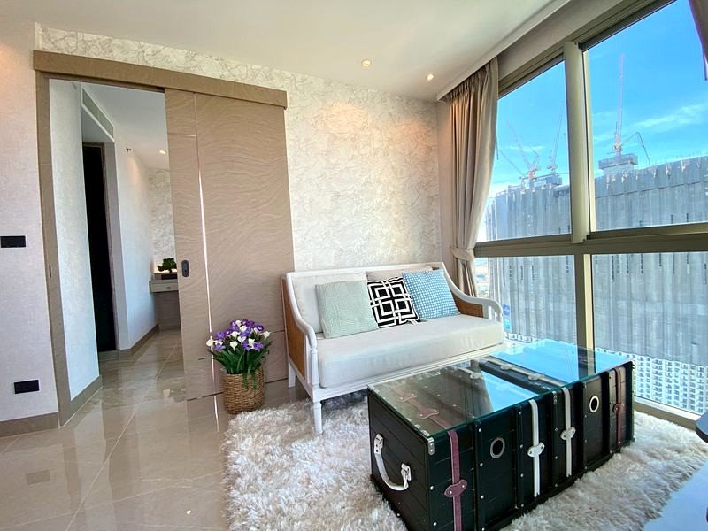 Bright living room with a white sofa, decorative pillows, a glass-topped trunk coffee table, and large windows showing a city view (cranes/skyline).
