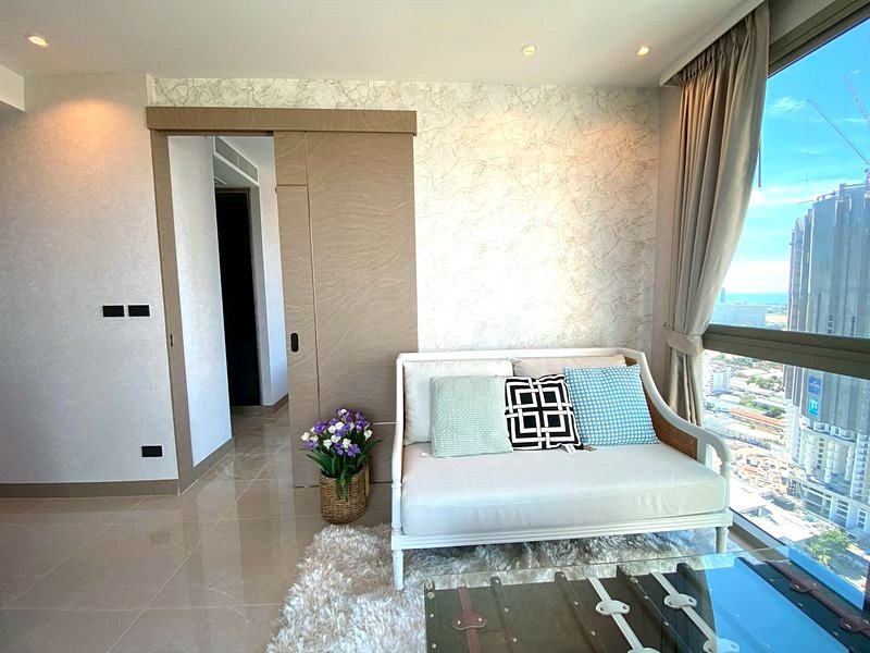 Bright modern living room with a white sofa and pastel cushions, sunlit from a large city-view window.