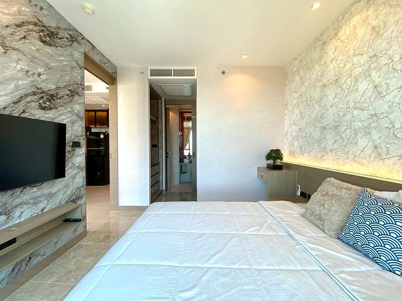 Hotel bedroom with a large bed, marble feature wall, and mounted flat-screen TV on the left; doorway to a hallway ahead.