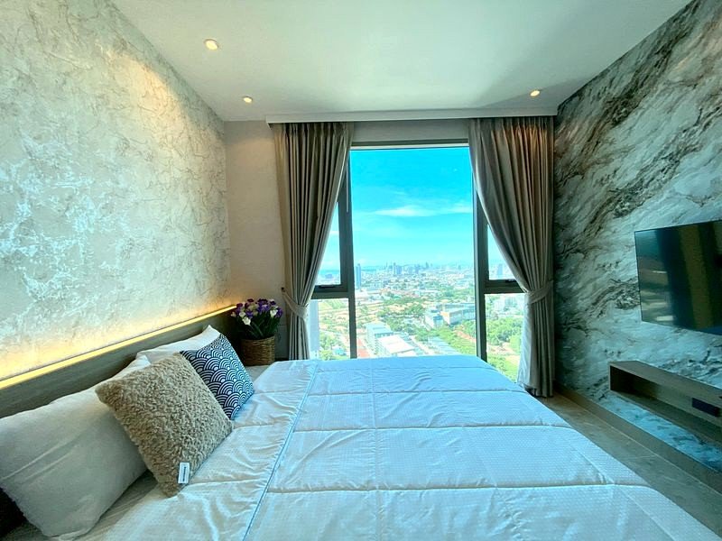 Hotel room with a large bed facing a floor-to-ceiling window showing a city skyline.