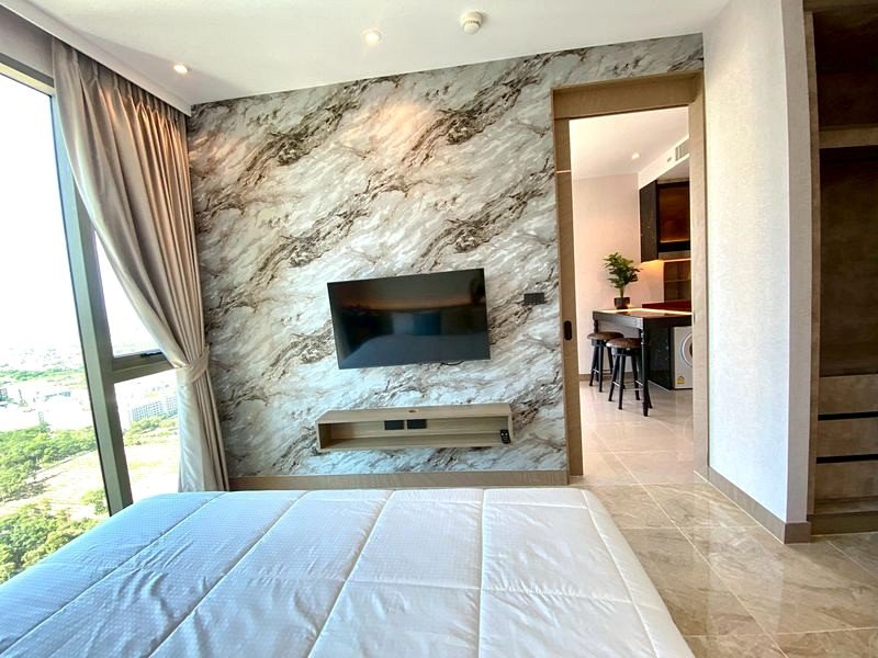 Bright hotel bedroom with a marble feature wall, wall-mounted TV, and a floating media shelf seen from the bed, plus a doorway to a kitchenette.