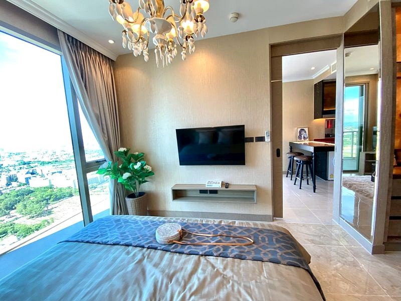 Hotel bedroom with a king bed, chandeliers, and a floor-to-ceiling window showing a city view; TV on the wall and a plant nearby.