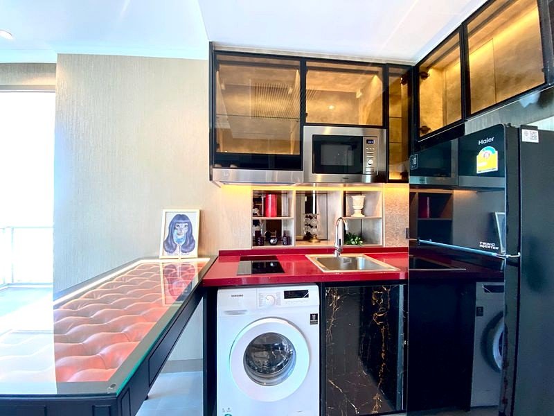 Modern, compact kitchen with red countertop, black cabinets, and built-in appliances (microwave, fridge) in an open-plan space, plus a washing machine under the counter.
