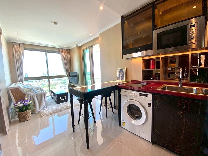 Open-plan kitchen and living area with a black glass-top bar, stools, and a washing machine beneath the counter by a large window.
