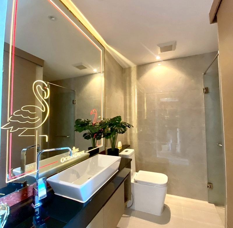 Modern bathroom with a backlit neon flamingo framed mirror, white vessel sink on a dark countertop, and potted plants.