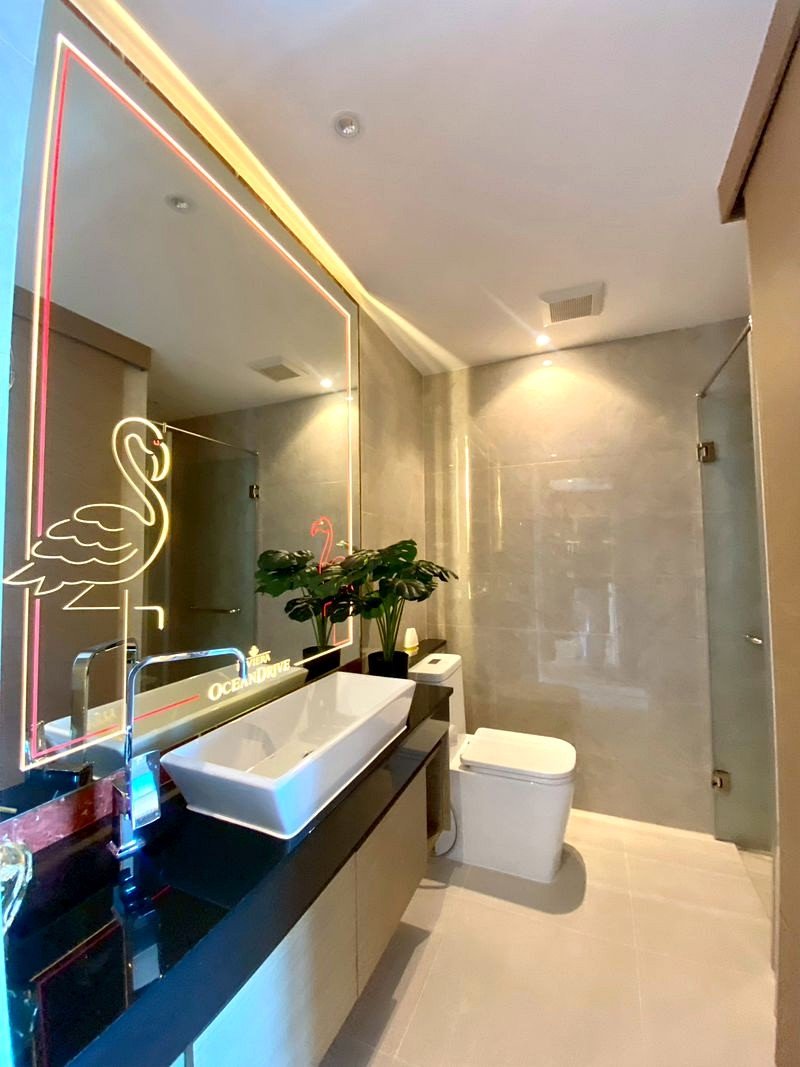 The_Riviera_Ocean_Drive_3914_16 Modern bathroom with a backlit neon flamingo framed mirror, white vessel sink on a dark countertop, and potted plants.