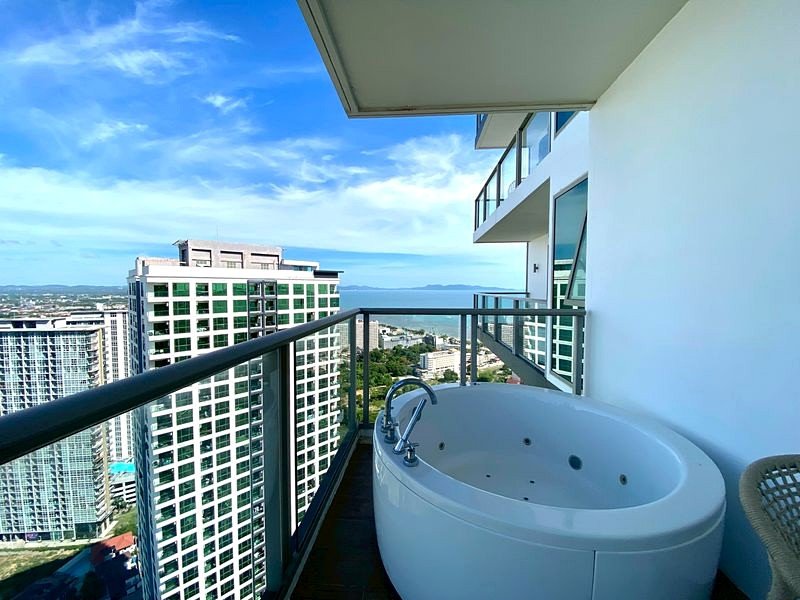 White freestanding jetted tub on a high-rise balcony with glass railing, overlooking city buildings and the ocean under a blue sky.