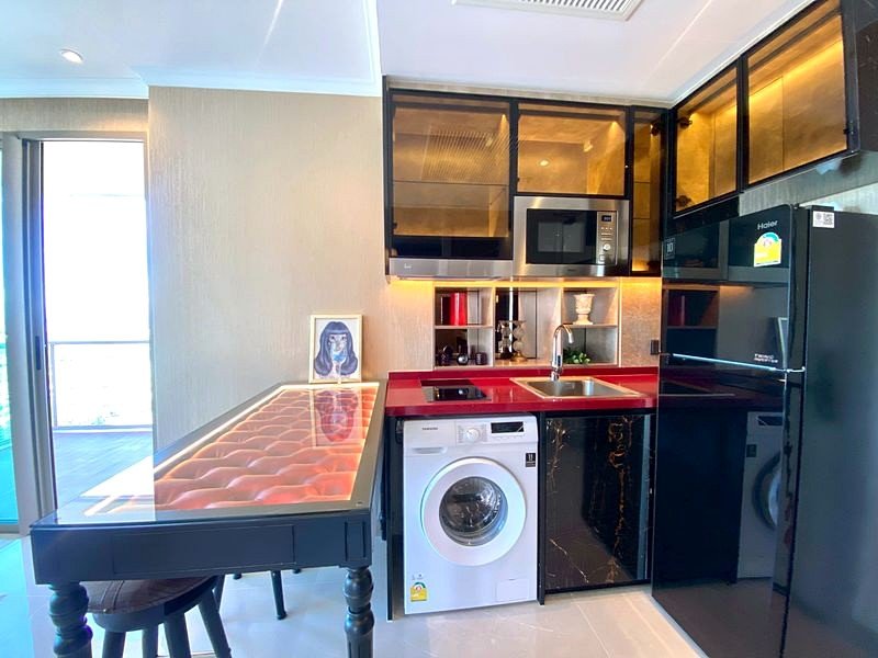 Compact modern kitchen with red counters, black gloss cabinets, a washing machine, microwave, and a glass-topped dining bar.