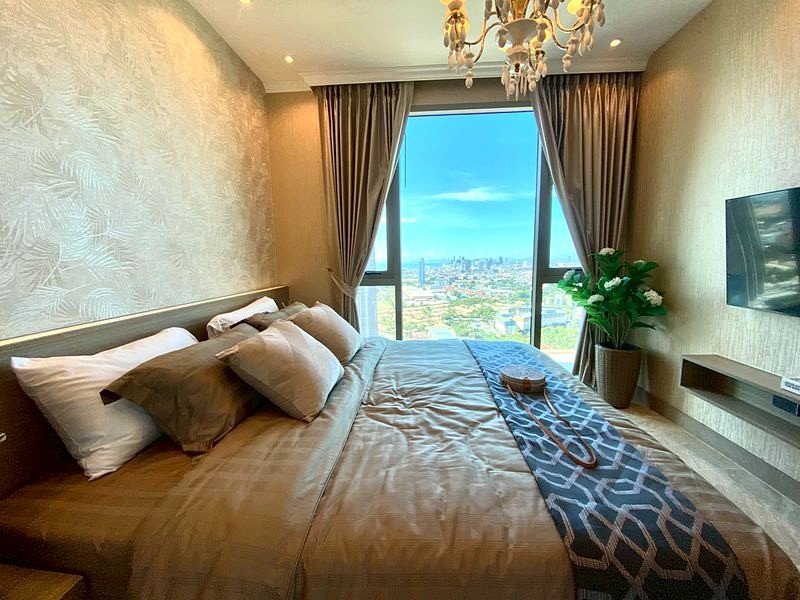 Luxurious hotel bedroom with a king bed, brown bedding and decorative pillows, and a city view through large windows.