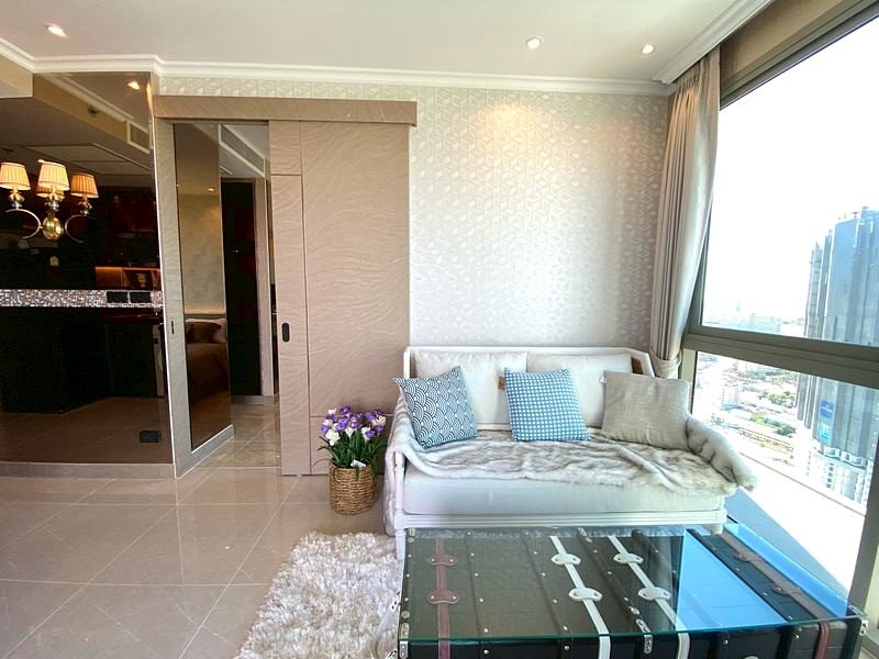 Bright living room with a white sofa, blue cushions, beige throw, and a glass coffee table overlooking the city skyline through a large window.