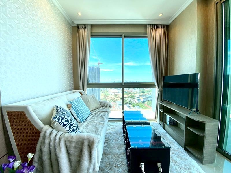 Modern living room with a white sofa, blue patterned cushions, and a glass coffee table facing a large window with a city view.