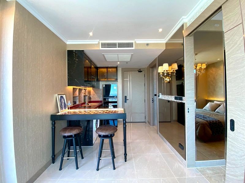 Open-plan hotel room with a small bar-height table and two stools, a compact kitchenette along the left, and a doorway to a separate bedroom visible through mirrored panels in the hallway.