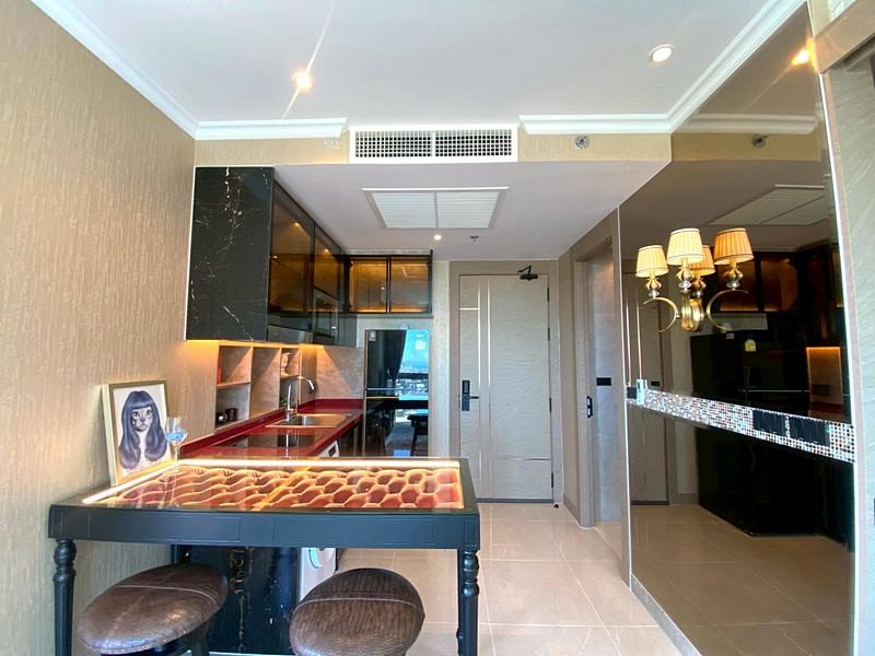 A modern kitchenette with a tufted leather bar table and two stools, black cabinets, red countertops, and warm lighting in a hotel-style suite.