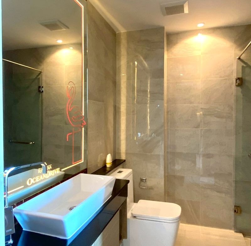 Modern bathroom with a white vessel sink on a dark countertop, large backlit mirror featuring a pink neon flamingo, toilet, and glass shower.