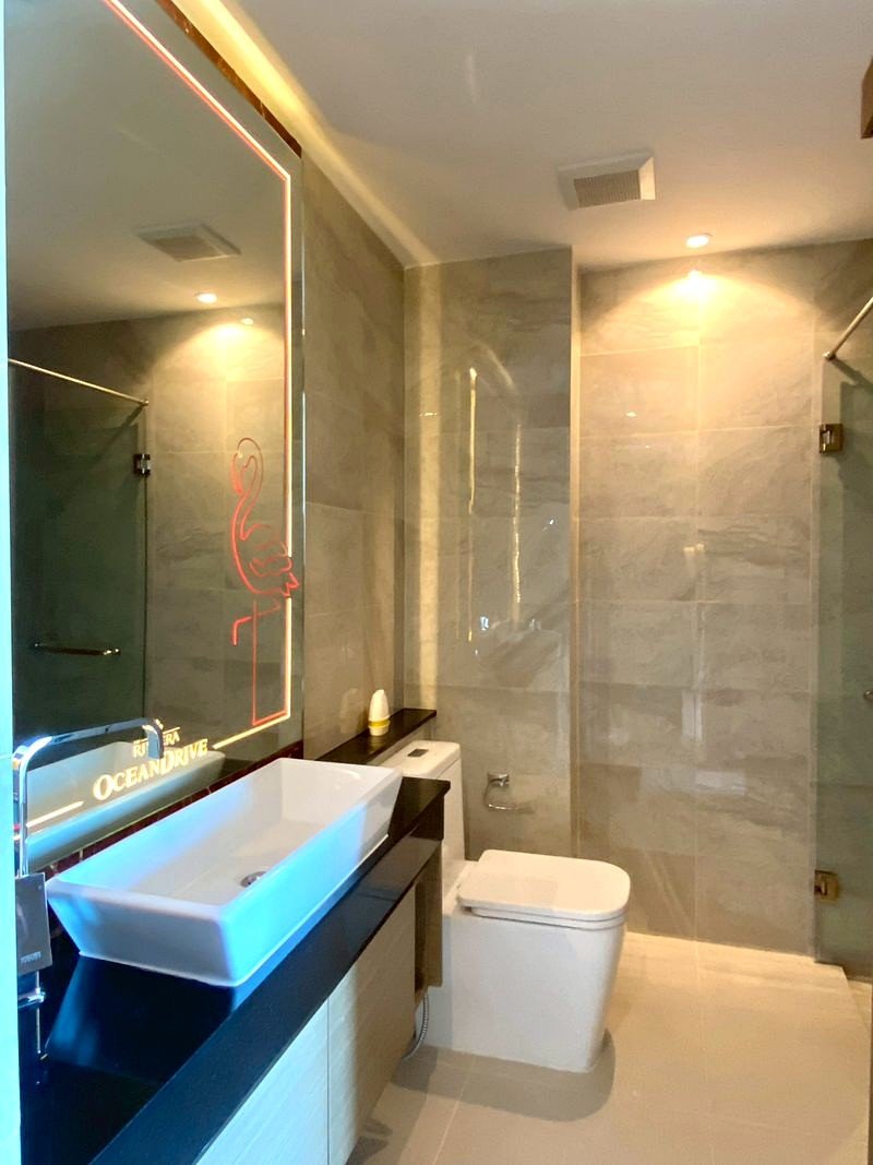 The_Riviera_Ocean_Drive_705_11 Modern bathroom with a white vessel sink on a dark countertop, large backlit mirror featuring a pink neon flamingo, toilet, and glass shower.