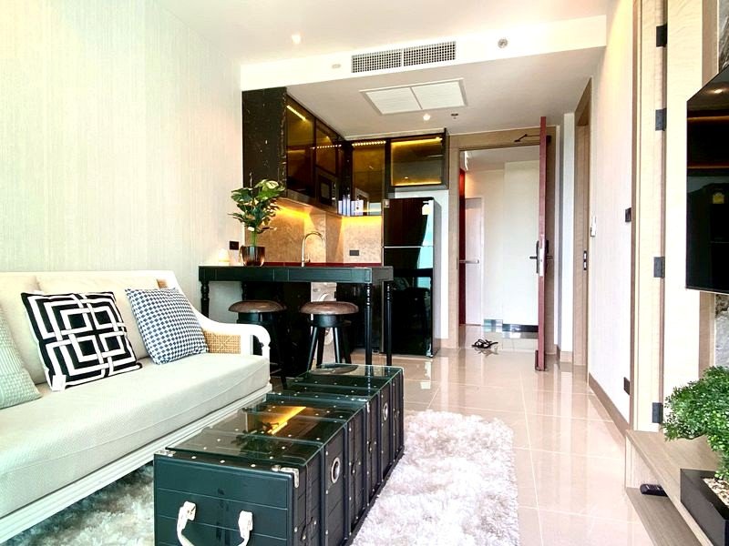 Open-concept living area with a pale sofa, geometric cushions, a dark green kitchen bar, and two bar stools.