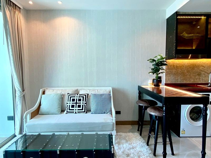Bright open living area with a white sofa, patterned cushions, and a glass-topped coffee table beside a window with curtains.