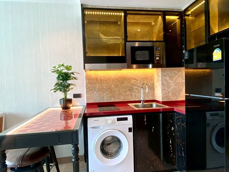 Compact modern kitchen with red countertops, sink, and built-in appliances including oven, microwave, and washing machine beneath the counter.