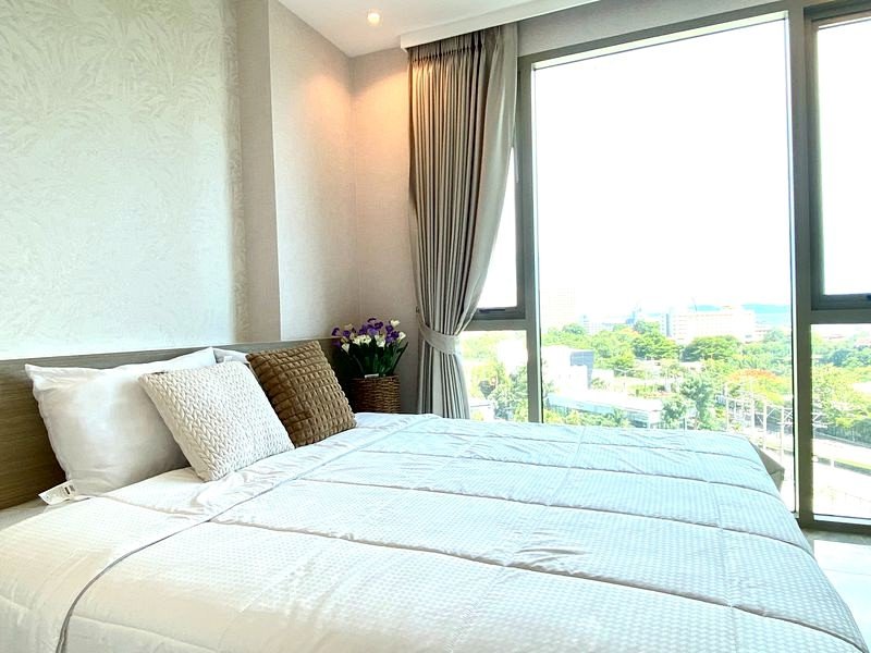 Hotel bedroom with white linens, decorative pillows, a vase of purple flowers, and a large window with a city view.