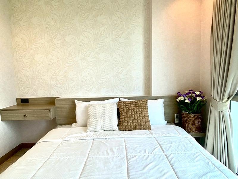 Hotel room with a neatly made bed, white bedding and two decorative pillows; a nightstand with a basket of flowers on the right.