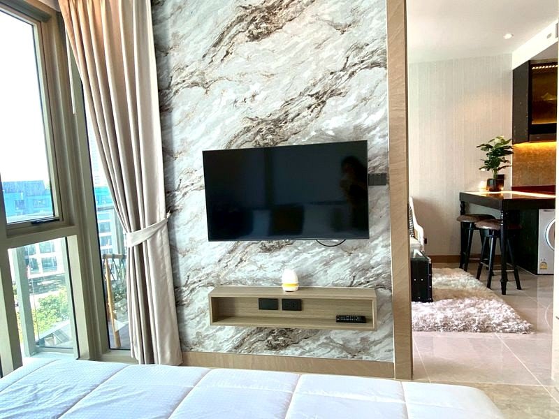 Hotel suite living area with a wall-mounted flat-screen TV on a marble accent wall, a wooden media shelf, and beige curtains by a large window.