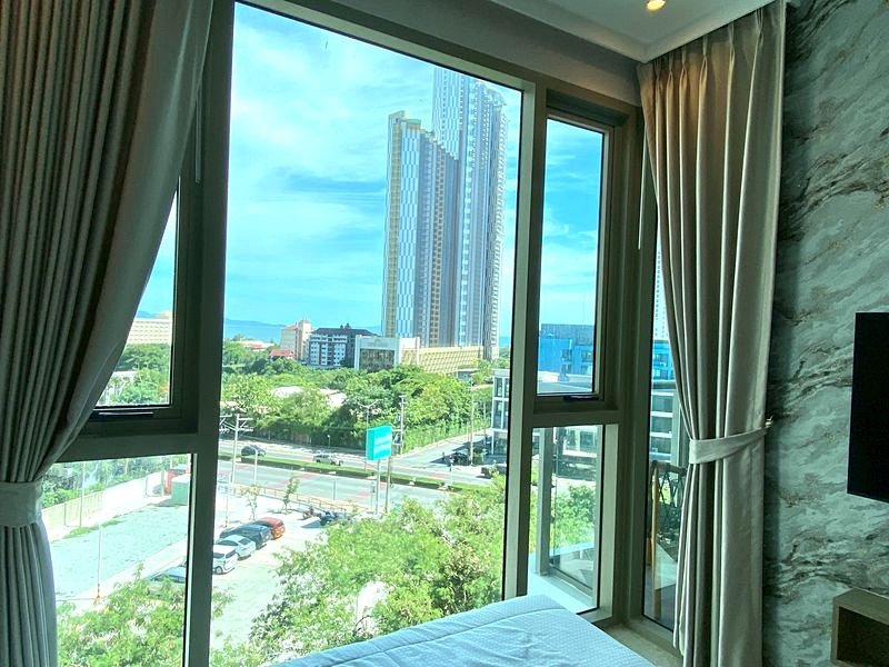 Hotel room with large windows and beige curtains overlooking a city skyline, including a tall modern skyscraper and a tree-lined street below, daytime view.
