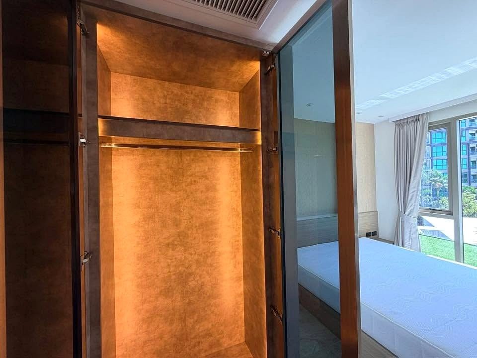 Open wardrobe with warm lighting beside a glass wall; visible bedroom with a bed and large windows.
