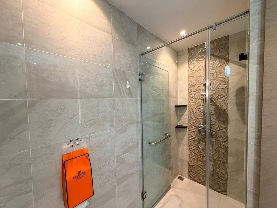 Glass-walled shower enclosure in a modern bathroom with beige tiles, a patterned tiled wall inside the shower, and built-in shelves; an orange shopping bag hangs on the wall nearby.
