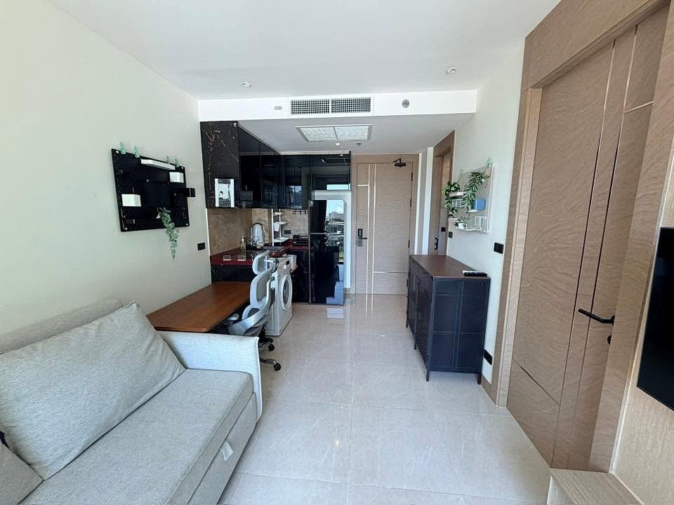 Bright open studio with a gray sofa, wooden desk, and white office chair near a black glossy kitchen area.