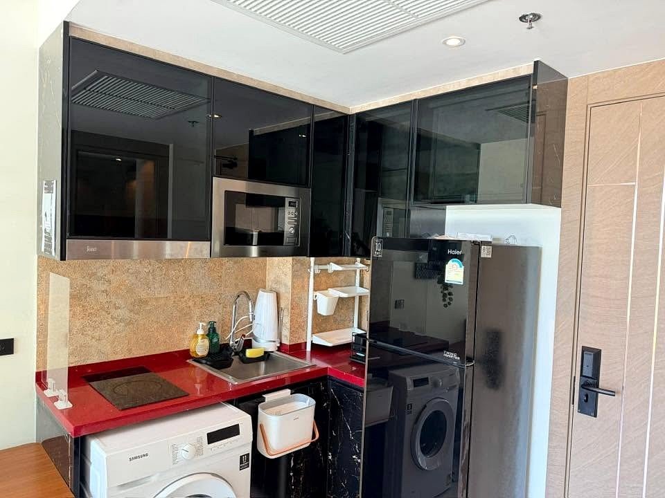 Compact kitchenette with red countertop, black glossy upper cabinets, sink, washing machine, microwave, and a Haier fridge.