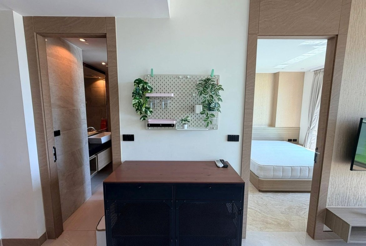 Modern hotel suite living area with pegboard wall, green plants, and a dark wooden island; bed visible in the background.