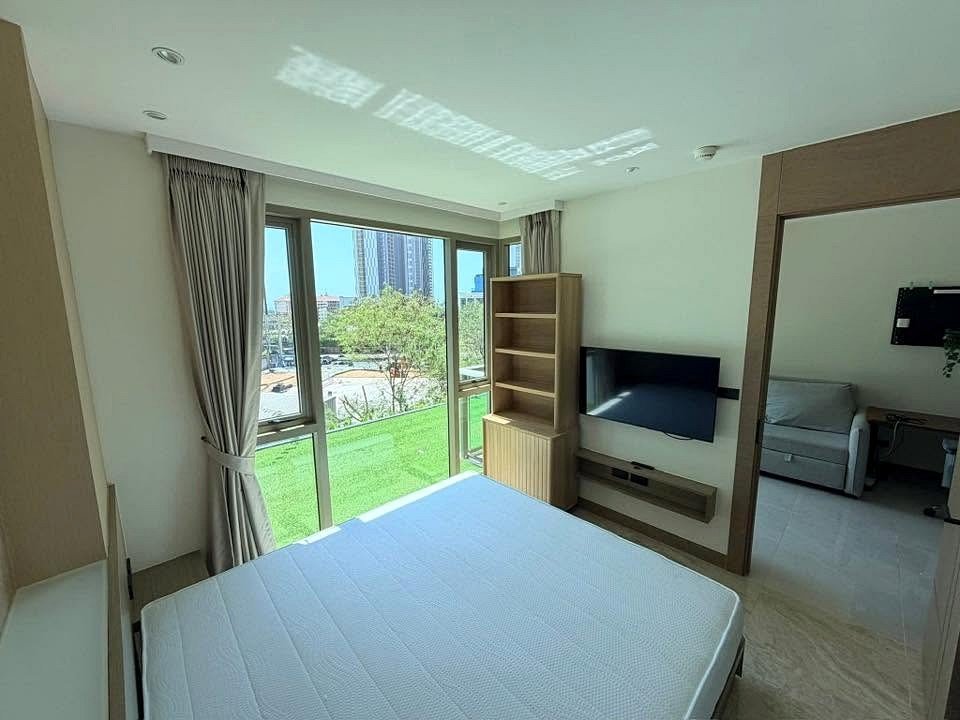 Hotel room with a large window and a bed, TV mounted on the wall, and a wooden shelving unit facing the window view of buildings and green outdoor space, through a doorway to a living area.
