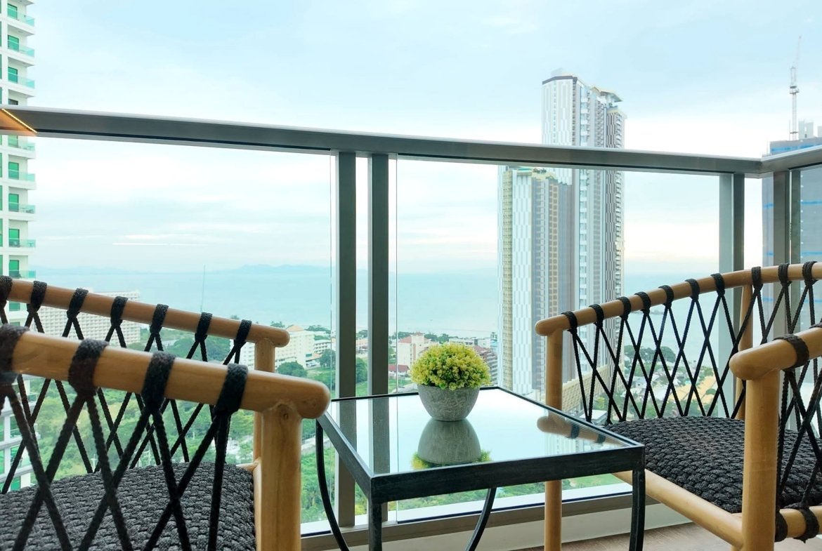 Balcony seating area with a glass table and potted plant, rope-woven bamboo railing, and a city skyline beyond.