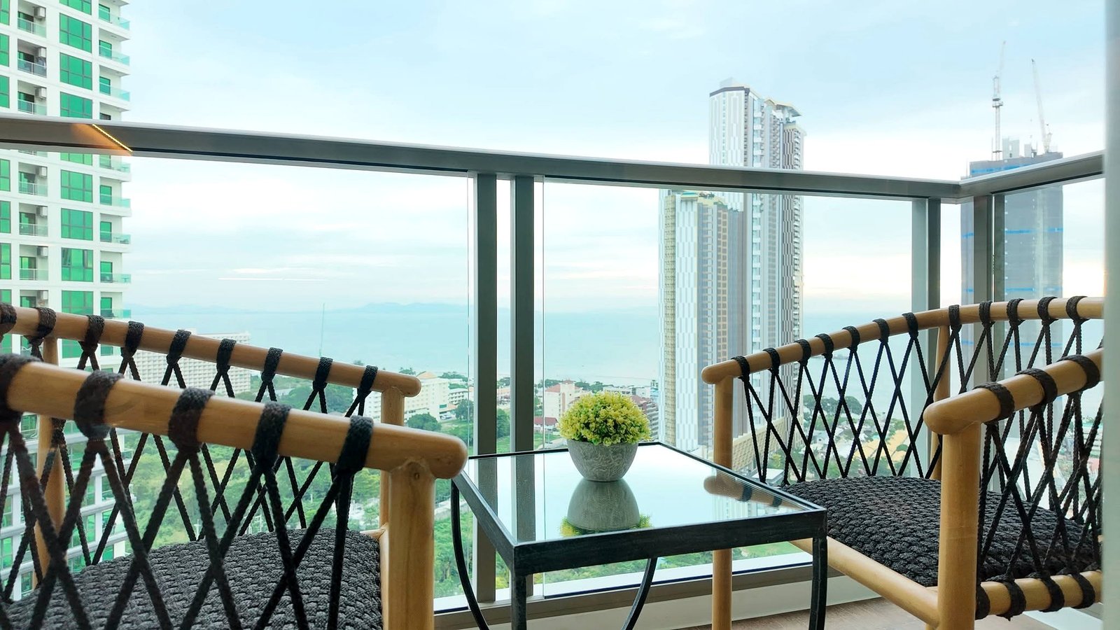 The_Riviera_Ocean_Drive_Pattaya_Dlc601_15 Balcony seating area with a glass table and potted plant, rope-woven bamboo railing, and a city skyline beyond.