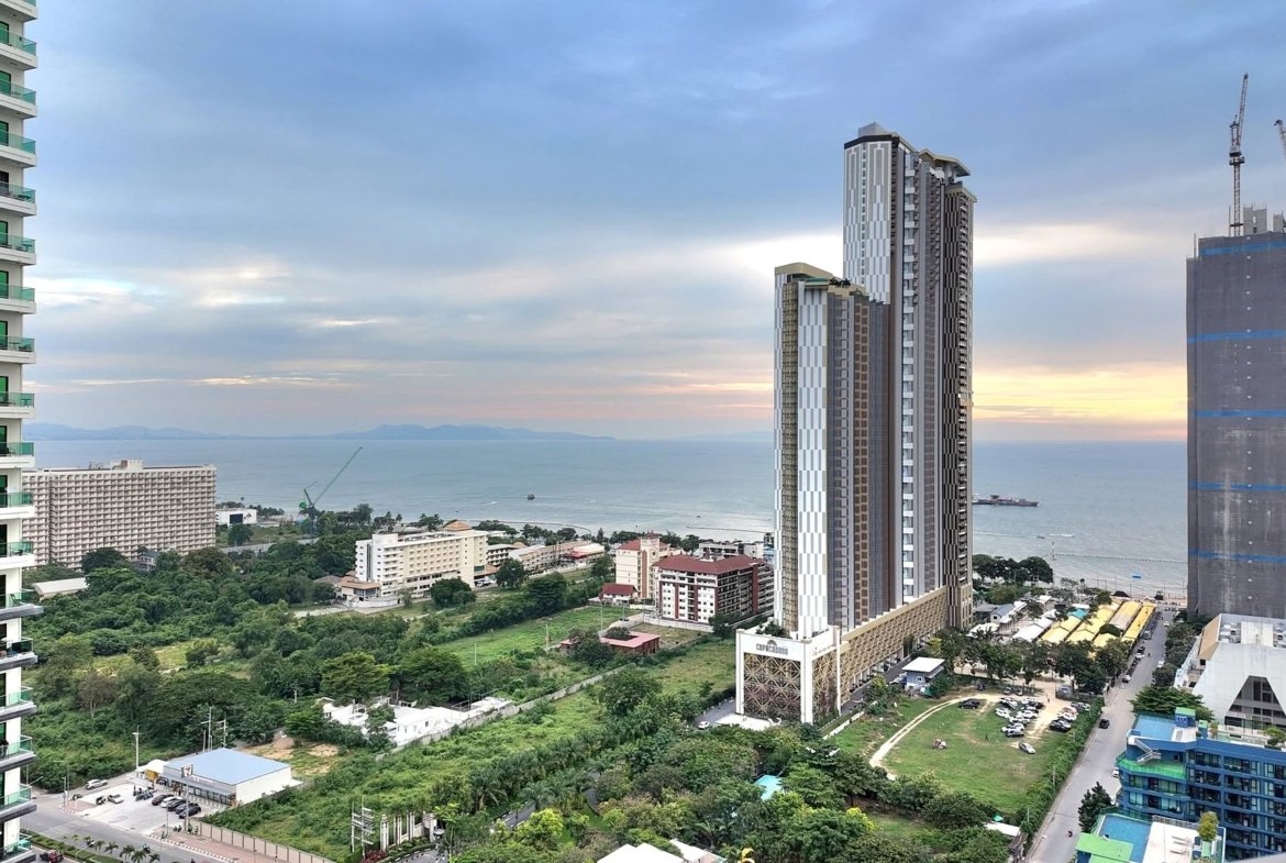 Modern city skyline by the sea: a green-glass residential tower on the left and tall high-rises with cranes on the right, under a cloudy sky.