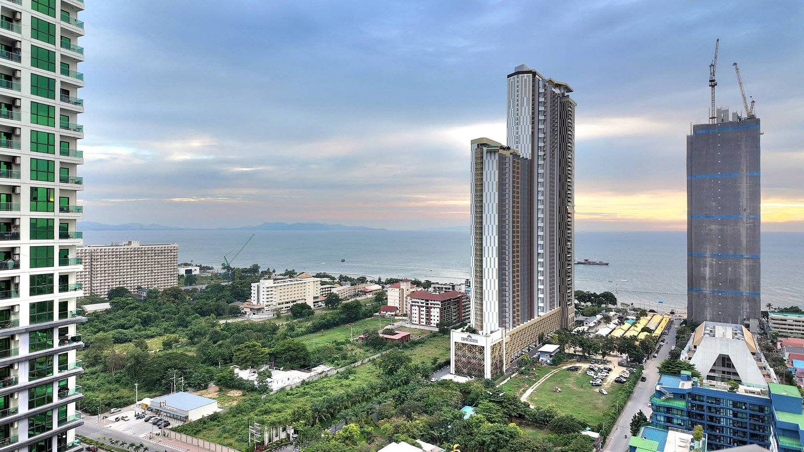 The_Riviera_Ocean_Drive_Pattaya_Dlc601_16 Modern city skyline by the sea: a green-glass residential tower on the left and tall high-rises with cranes on the right, under a cloudy sky.