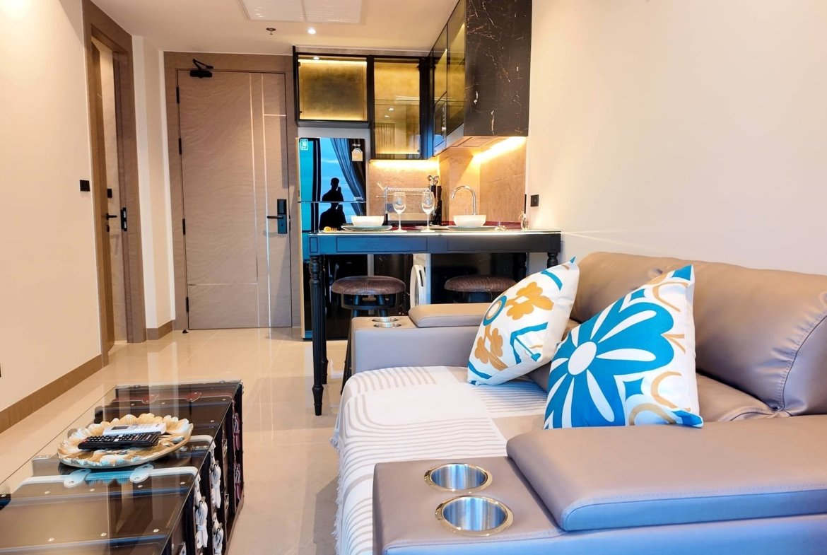 Open‑concept living area with a beige sofa and blue/yellow floral pillows, a glass coffee table, and a small kitchen behind it.