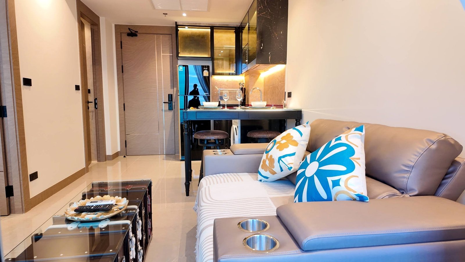 The_Riviera_Ocean_Drive_Pattaya_Dlc601_2 Open‑concept living area with a beige sofa and blue/yellow floral pillows, a glass coffee table, and a small kitchen behind it.