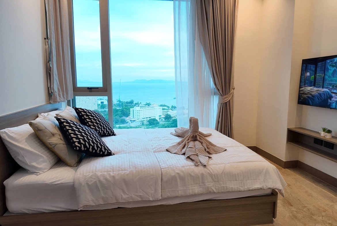 Hotel bedroom with large window view of the ocean and distant mountains, sheer and blackout curtains drawn to the side.