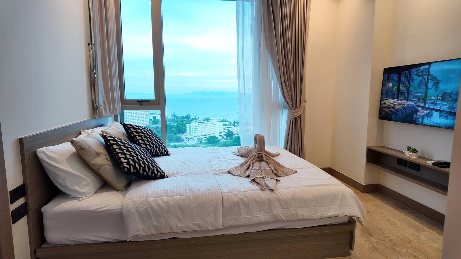 The_Riviera_Ocean_Drive_Pattaya_Dlc601_3 Hotel bedroom with large window view of the ocean and distant mountains, sheer and blackout curtains drawn to the side.