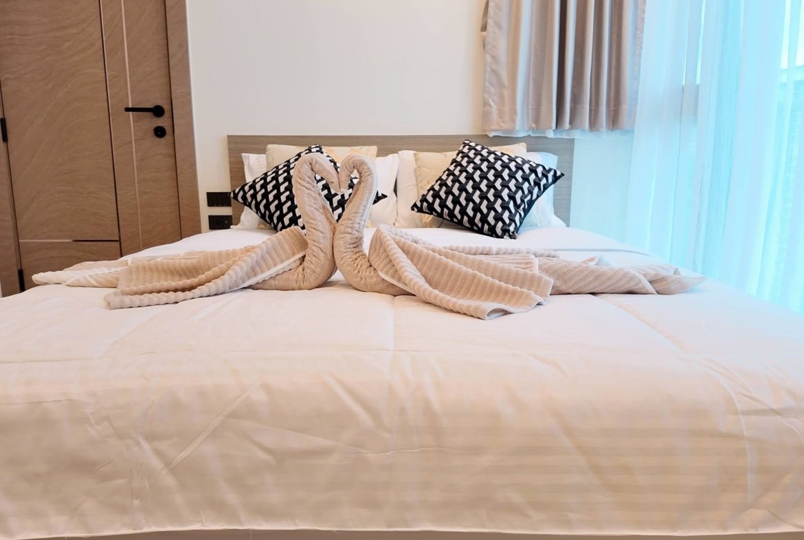 Bed with white bedding and two decorative pillows; beige towels folded into swans form a heart in the center.