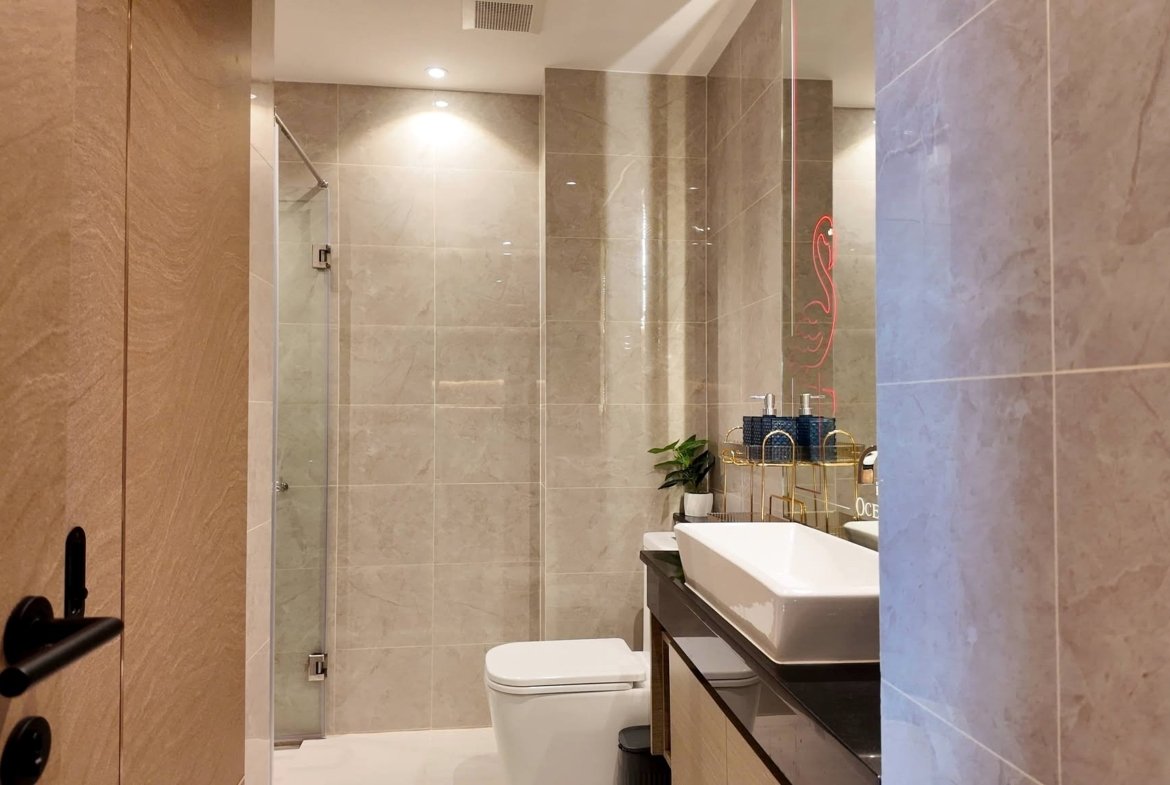 Modern bathroom with beige marble tiles, glass shower, white basin, and a sleek dark vanity; a potted plant sits by the sink.